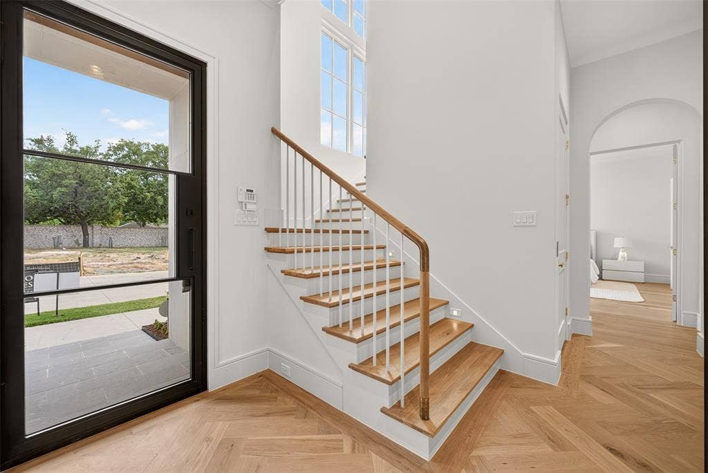 Herringbone wood-finish flooring, staircase with wood treads and white risers, black-framed glass entry door, white walls, and an arched hallway entry
