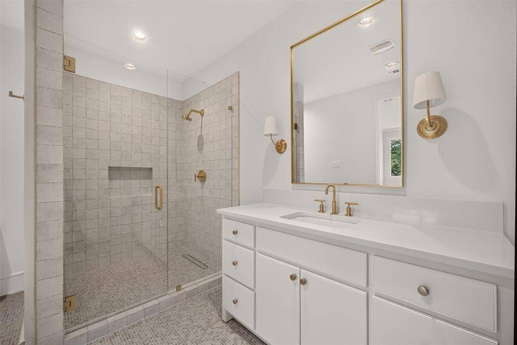 Frameless glass shower enclosure with tiled walls and floor, single vanity with white cabinetry, integrated sink, and gold-finish fixtures and mirror