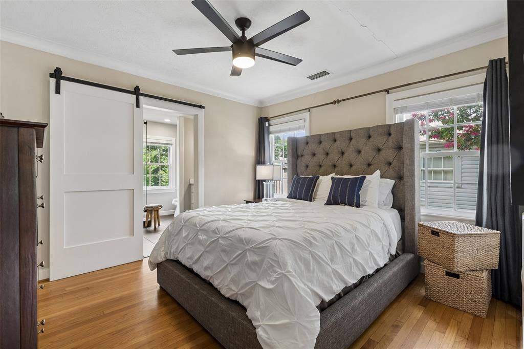 Hardwood flooring, white barn door, ceiling fan with light, crown molding, and multiple windows