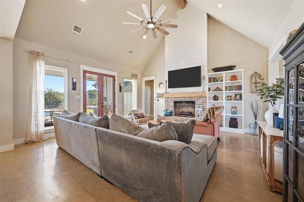 Living room featuring french doors, high vaulted ceiling, a brick fireplace, a ceiling fan, and finished concrete floors