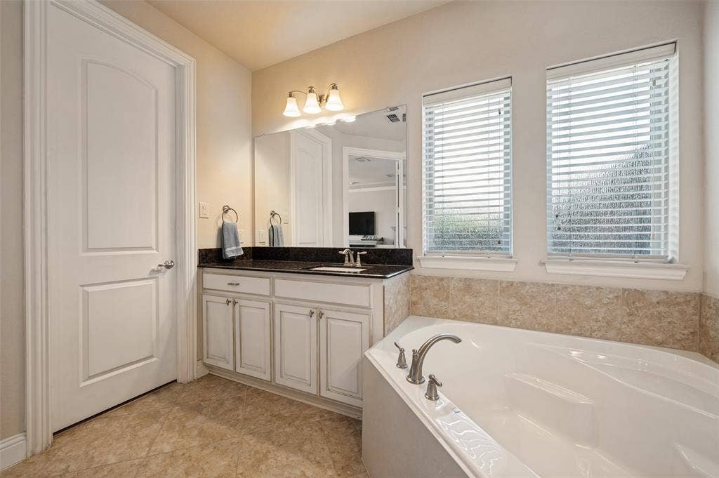 741 Darian Drive, Prosper, TX 75078 Soaking tub with brushed nickel fixtures, dual windows with blinds, single vanity with dark countertop, light-colored cabinetry, and tile flooring