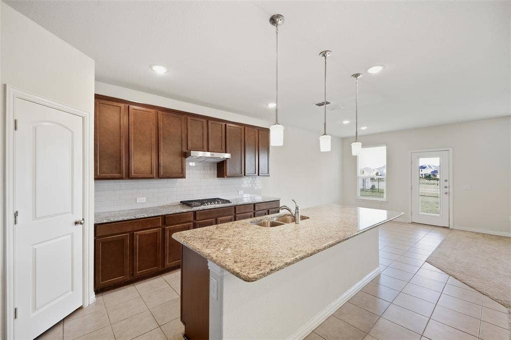 1805 Nelson Street, Anna, TX 75409 Kitchen featuring wood cabinetry, a granite-finish island with a double basin sink, stainless steel gas cooktop, tile backsplash, and pendant lighting