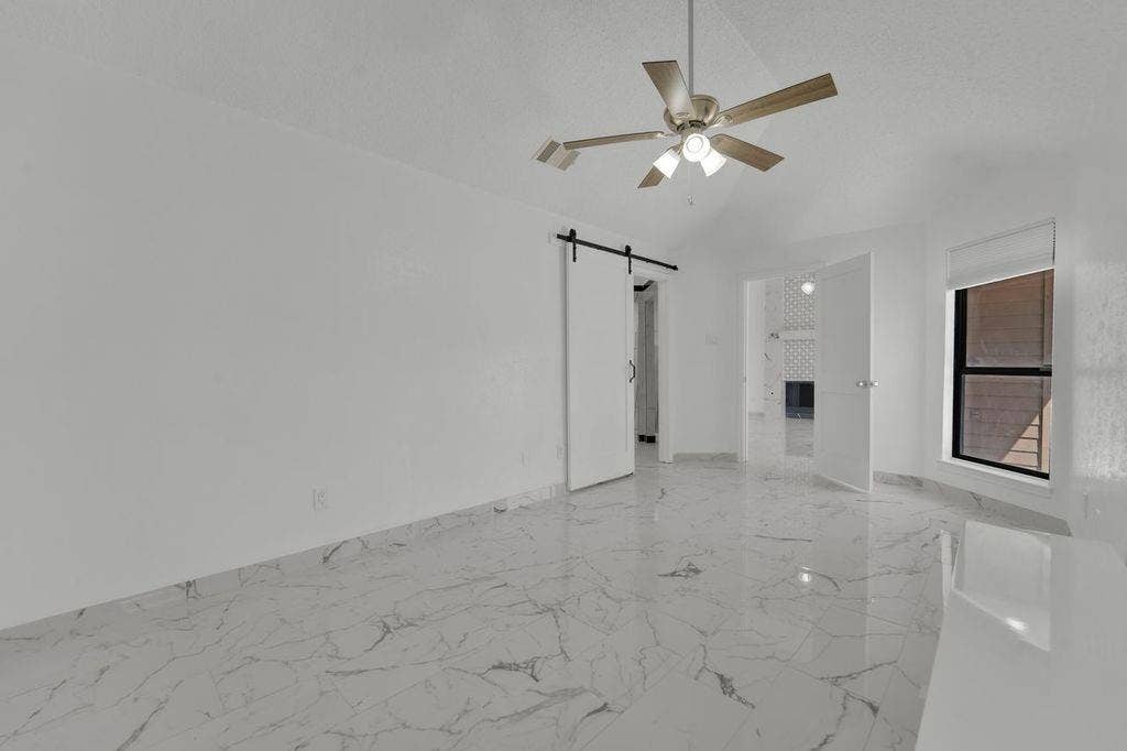 Vaulted ceiling with ceiling fan, white walls, marble-look tile flooring, barn door, and a large window
