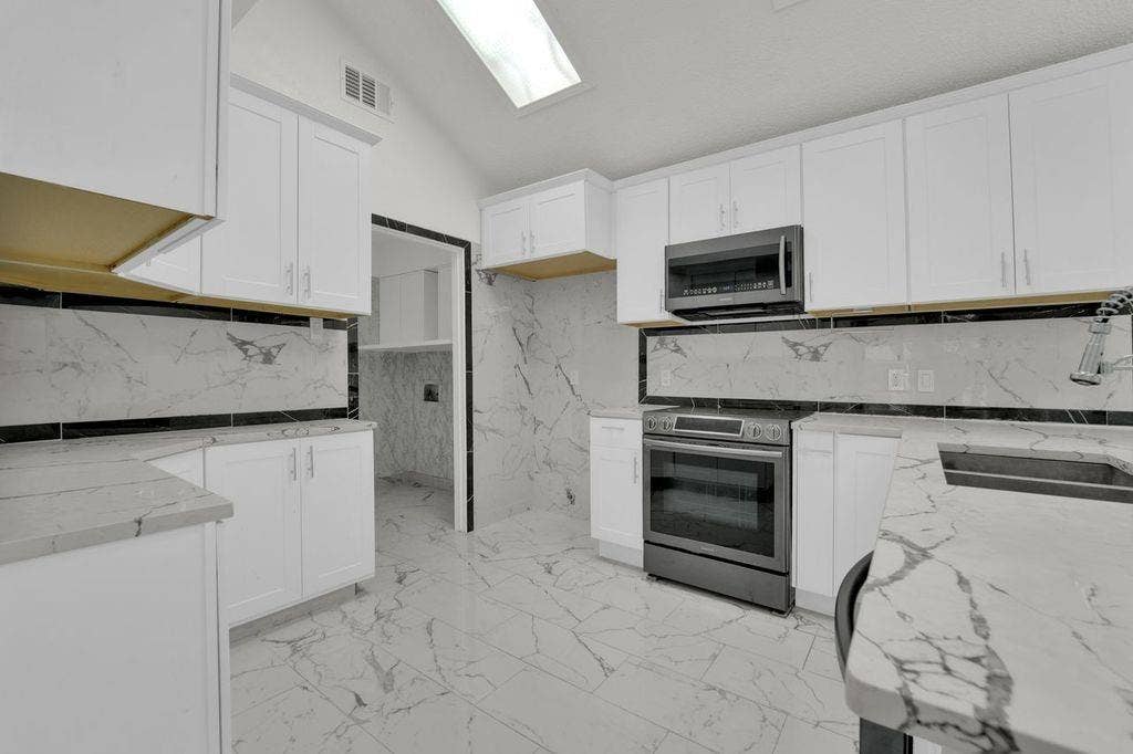 Vaulted ceiling with skylight, white shaker cabinetry, stainless steel oven, built-in microwave, and marble-look countertops with integrated sink