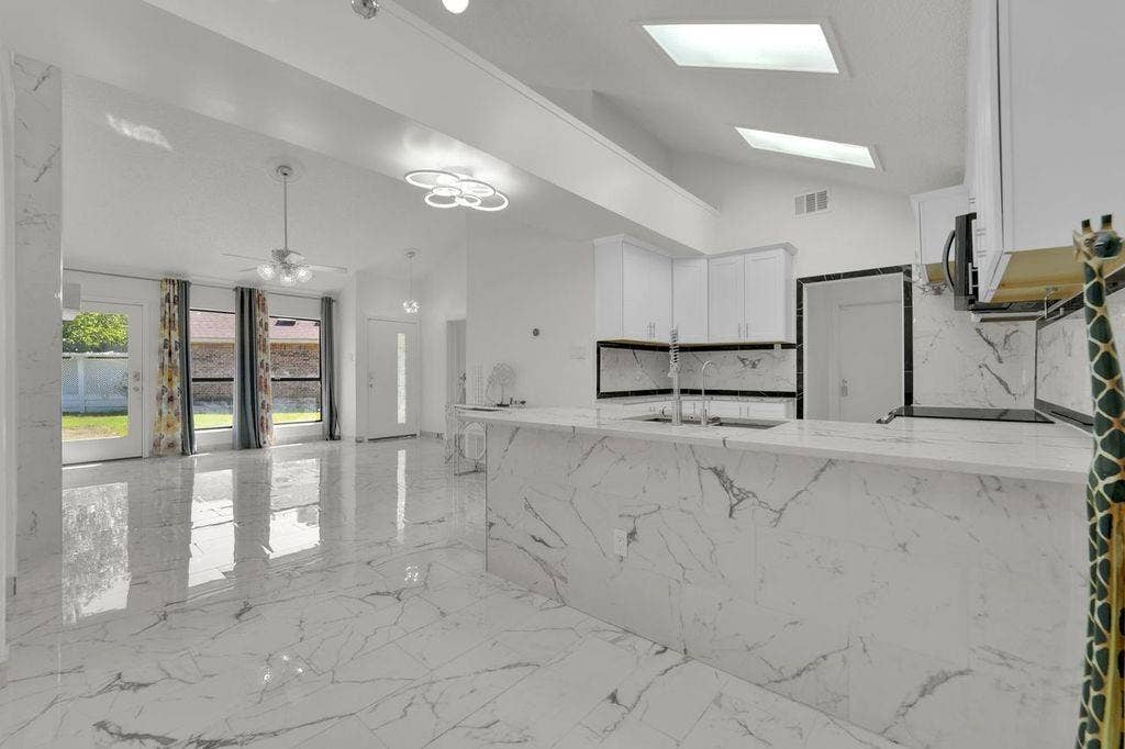 Vaulted ceilings with skylights, white cabinetry, a kitchen island with an integrated sink, and white marble-look flooring
