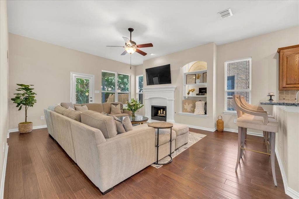 5612 Hampshire Drive, McKinney, TX 75070 Wood-finish flooring throughout, vaulted ceiling with a ceiling fan, fireplace with a mantel, built-in shelving, and a breakfast bar with stone countertops