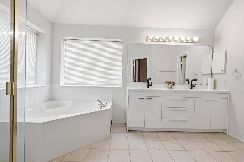Corner soaking tub, dual vanity with white cabinetry, tile flooring, and a large wall-mounted mirror