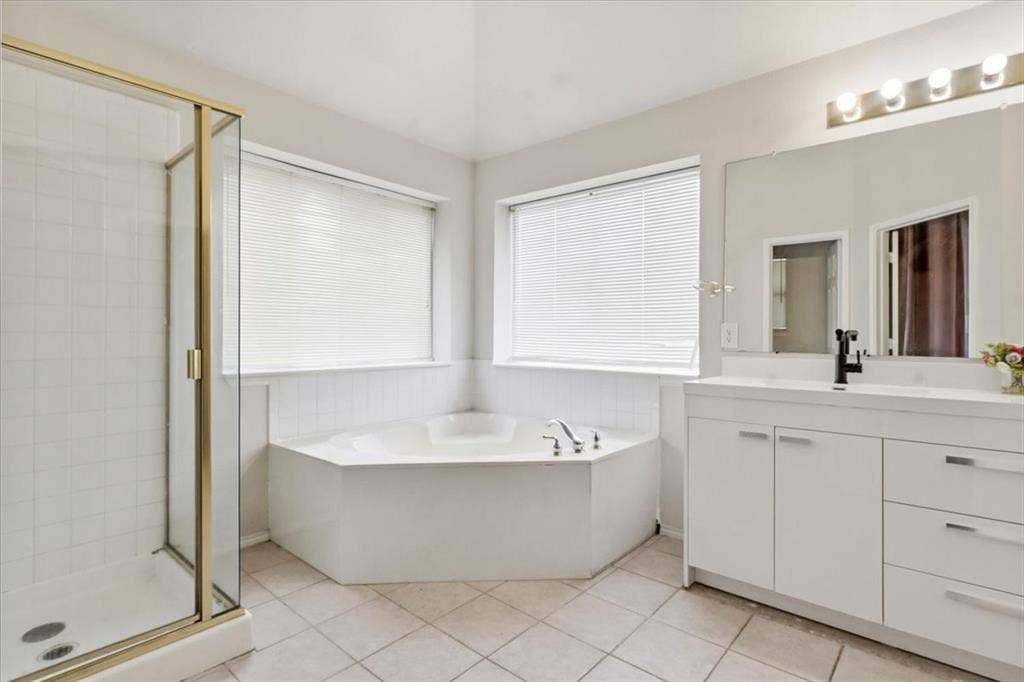 Corner soaking tub, glass-enclosed shower, dual-sink vanity with white cabinetry, wall-mounted vanity lighting, and tiled flooring