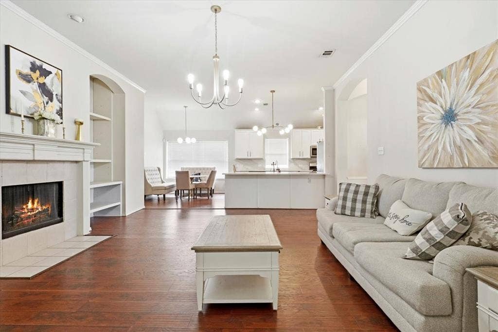 Wood-finish flooring, white fireplace with tile surround, built-in shelving, white kitchen cabinetry, and overhead lighting fixtures