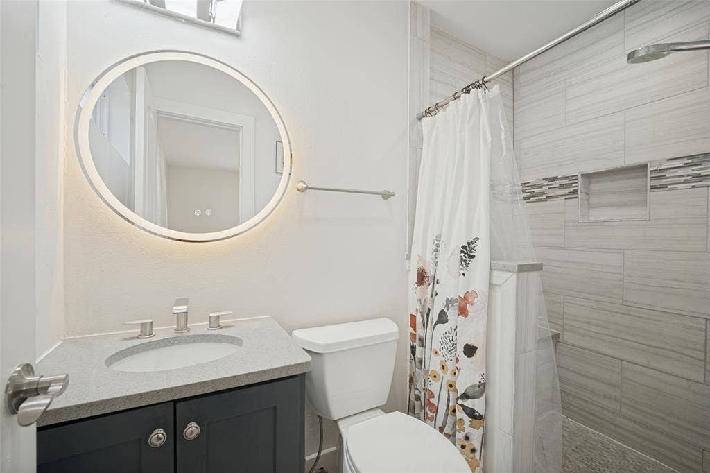 3209 Karla Drive, Mesquite, TX 75150 Bathroom featuring a round LED-backlit mirror, dark-finish vanity with an integrated sink, and a tiled shower with a mosaic accent band and built-in niche