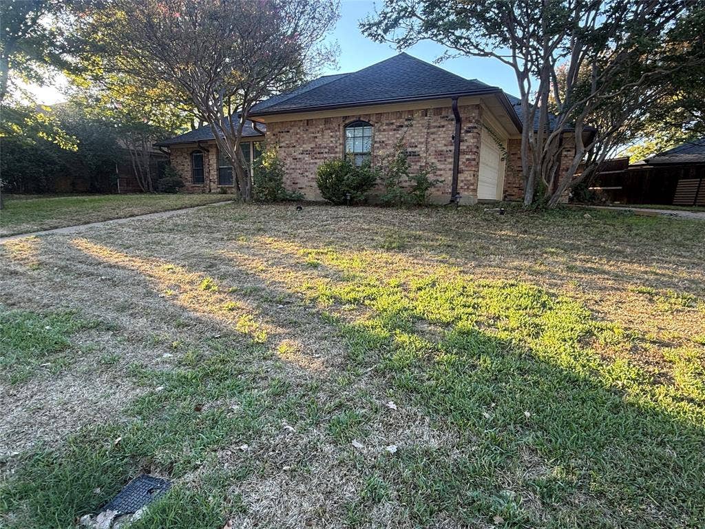 3407 Ruidoso Drive, Arlington, TX 76017 Brick exterior, dark shingle roof, attached garage, established trees, and front yard