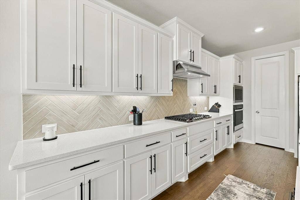 White shaker cabinetry, herringbone tile backsplash, stainless steel range hood, built-in double ovens, and wood-finish flooring
