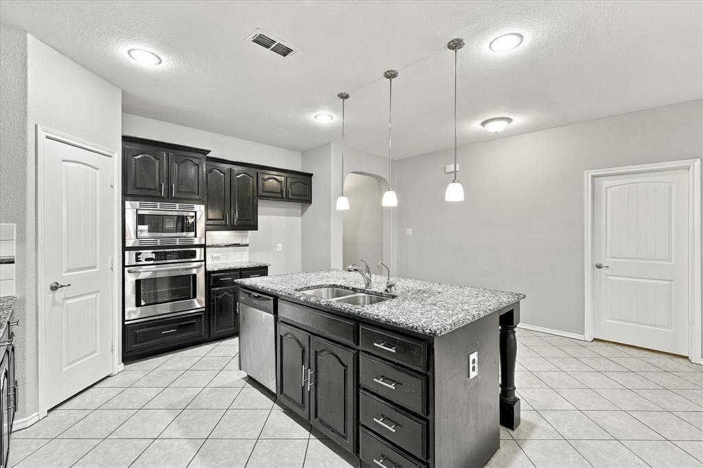 4716 Kendrick Peak Way, Fort Worth, TX 76244 Kitchen featuring dark wood cabinetry, stainless steel built-in appliances, a central island with a sink and granite-style countertop, and white tile flooring