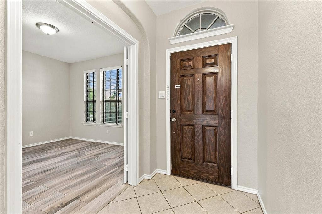 4716 Kendrick Peak Way, Fort Worth, TX 76244 Wood-paneled entry door with an arched transom window, ceramic tile flooring, and a recessed doorway leading to a room with wood-finish flooring and two windows with blinds