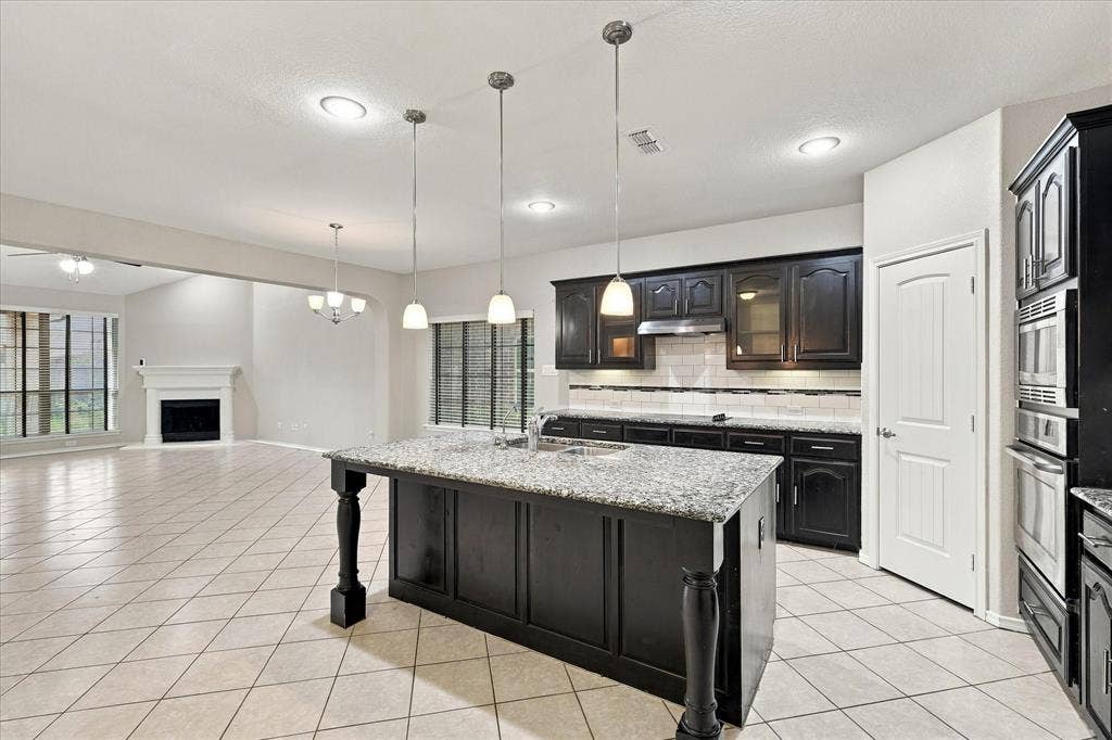 4716 Kendrick Peak Way, Fort Worth, TX 76244 Kitchen island with granite countertop and pendant lighting, dark wood cabinetry, tile flooring, built-in stainless steel appliances, and a white mantel fireplace in the adjacent room