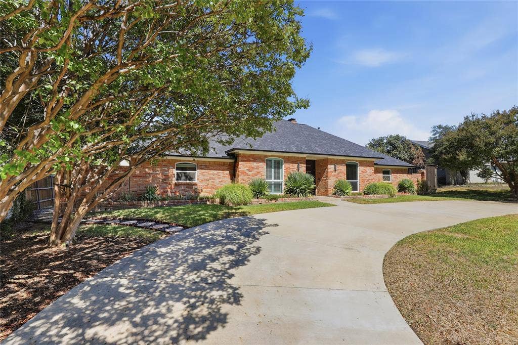 Brick exterior with a dark shingle roof, mature landscaping, and an expansive concrete driveway