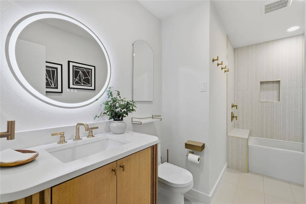 Backlit circular mirror, a single vanity with stone countertop, and a built-in bathtub with tiled surround and niche