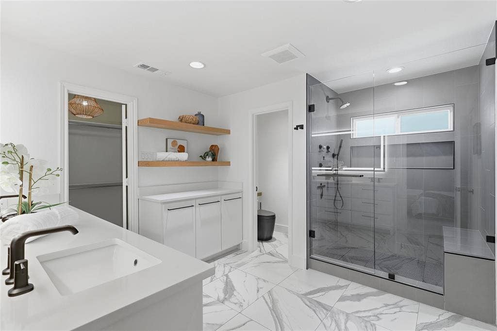 Walk-in shower with clear glass enclosure and integrated bench, wood-finish floating shelves, recessed lighting, and a single vanity with an undermount sink and dark bronze faucet