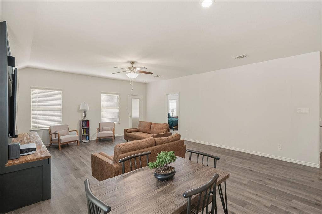 202 Buckboard Lane, Caddo Mills, TX 75135 The main living area inside 202 Buckboard Ln showcases a bright, open-concept floor plan designed for effortless daily living. Durable faux-wood luxury vinyl plank flooring runs beneath neutral off-white walls, seamlessly unifying the dining space and living room. Double-hung windows with faux-wood blinds and a glass-inset exterior door along the back wall fill the interior with daylight. Modern recessed lights and a ceiling fan complete the single-story layout, which features clear sightlines toward an adjoining bedroom wing for practical everyday convenience.