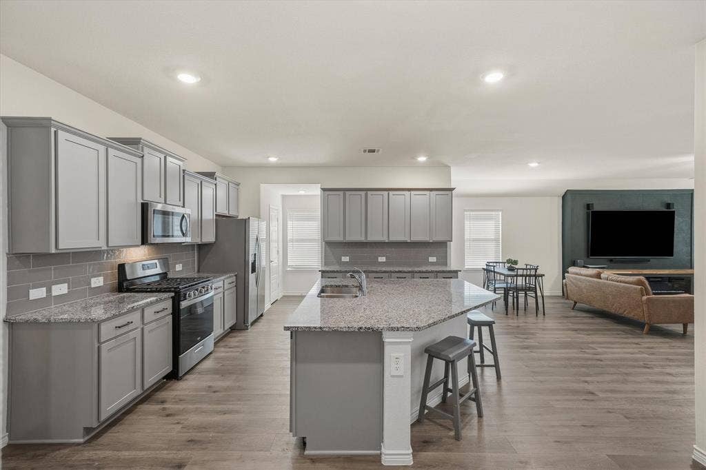 202 Buckboard Lane, Caddo Mills, TX 75135 This open-concept interior at 202 Buckboard Ln highlights seamless entertaining with durable gray wood-look plank flooring connecting the kitchen, dining, and living zones. The modern kitchen boasts gray Shaker cabinetry, speckled granite countertops, a subway tile backsplash, stainless steel appliances, and an angled eat-in island with breakfast bar seating. Recessed lighting brightens the kitchen and adjacent dining area, which flows naturally into the spacious great room featuring a bold dark accent wall, creating a stylish, cohesive layout ideal for everyday modern living.