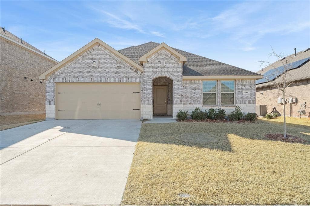 202 Buckboard Lane, Caddo Mills, TX 75135 Front exterior view of 202 Buckboard Ln highlights a charming single-story brick home beneath a clear blue sky. The facade combines light-toned masonry with a limestone water table, cross-gabled architectural rooflines, and a two-car front-entry garage with carriage-style hardware. An arched entry porch provides a welcoming focal point, complemented by tidy foundation shrubbery and a concrete driveway. Situated on a level suburban lot in Caddo Mills, this turnkey residence showcases balanced proportions and appealing curb appeal ideal for modern neighborhood living.