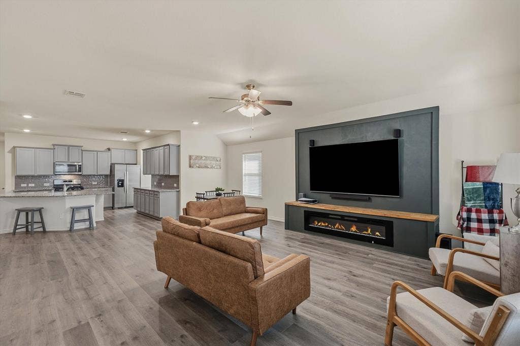 202 Buckboard Lane, Caddo Mills, TX 75135 The bright open-concept living space at 202 Buckboard Ln features modern wood-look laminate flooring that seamlessly connects the family room, dining nook, and kitchen. A striking dark accent media wall with a built-in linear electric fireplace anchors the living area beneath recessed LED lighting and a contemporary ceiling fan. In the background, the functional kitchen showcases painted shaker cabinets, granite countertops, and stainless steel appliances, creating an inviting layout ideal for relaxed everyday living and effortless indoor entertaining in Caddo Mills.