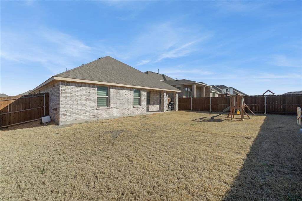 202 Buckboard Lane, Caddo Mills, TX 75135 A wide view captures the expansive, fully fenced backyard of the single-story home at 202 Buckboard Ln. Flat, level terrain stretches across the rectangular lawn, completely enclosed by a tall wooden privacy fence with sturdy metal posts. The rear exterior showcases light variegated brick, a covered back patio, and a poured concrete slab foundation beneath a low-pitch hip roof. Mid-afternoon sun illuminates the open outdoor space, which easily accommodates a detached wooden playset and swing set while leaving ample room for outdoor recreation and custom landscaping.