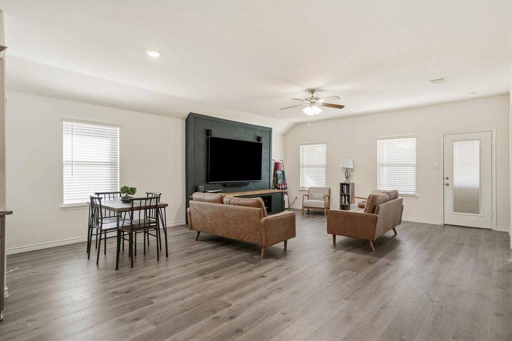 202 Buckboard Lane, Caddo Mills, TX 75135 The spacious open-concept living area at 202 Buckboard Ln offers a fluid, modern layout connecting the primary family room, dining space, and adjacent kitchen. Durable wood-look plank flooring extends beneath high ceilings equipped with recessed lighting and a classic ceiling fan. A deep hunter green accent wall serves as a central media focal point, while multiple single-hung windows fitted with blinds introduce soft natural light throughout. Direct exterior access via a frosted-glass door provides a seamless transition to the backyard, perfect for effortless indoor-outdoor Texas entertaining.
