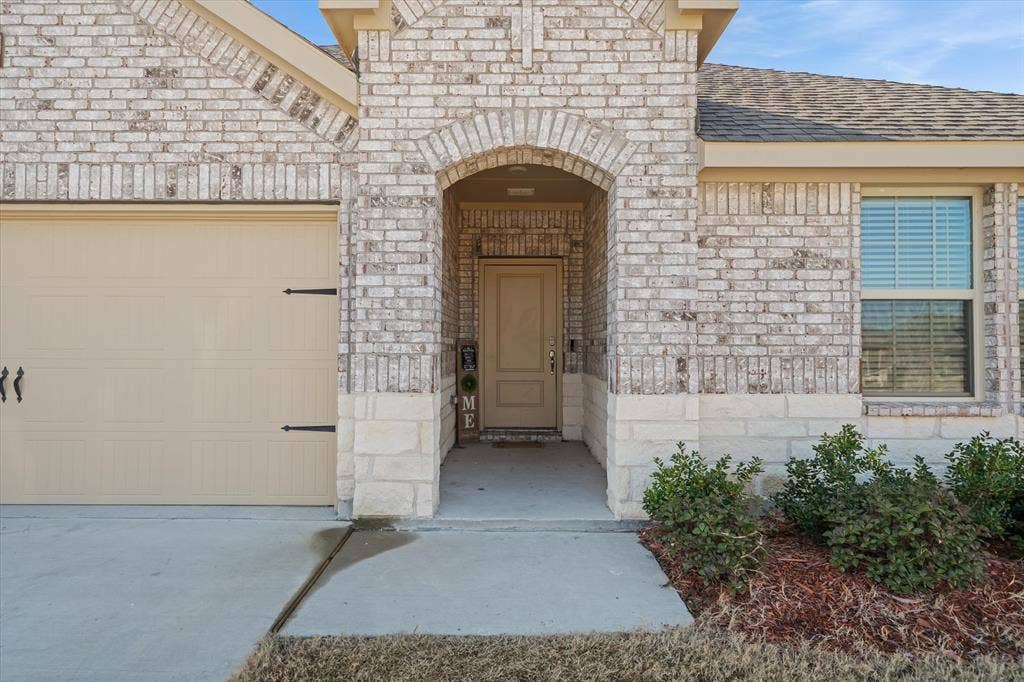 202 Buckboard Lane, Caddo Mills, TX 75135 Eye-level view of the front exterior at 202 Buckboard Ln, highlighting a modern single-story residence crafted with whitewashed brick and rough-cut limestone wainscoting. A graceful brick arch shelters the recessed entryway, complemented by a warm tan front door and a matching sectional garage door with decorative carriage-house hardware. A poured-concrete driveway and front walkway cross the level lawn, bordered by tidy red-mulch landscaping beds with evergreen shrubs, all presented beneath a clear sky in Caddo Mills.