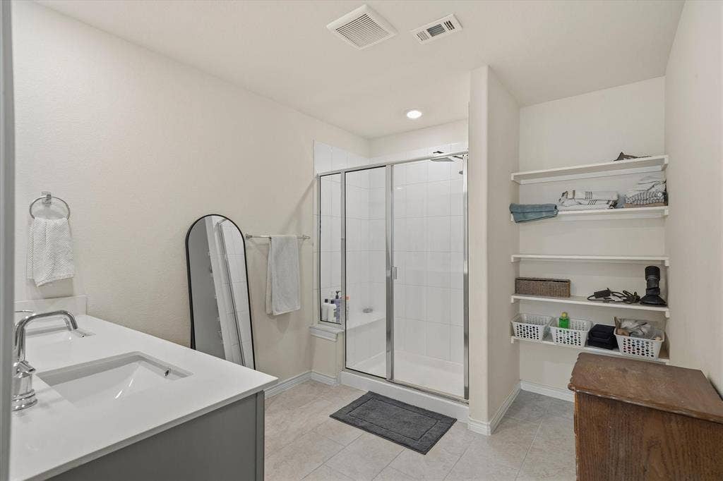 202 Buckboard Lane, Caddo Mills, TX 75135 Primary bathroom interior at 202 Buckboard Ln, Caddo Mills, showcasing neutral ceramic tile flooring and light gray cabinetry with a crisp white solid-surface countertop. The functional layout features a contemporary undermount sink with chrome fixtures, an arched black-framed floor mirror, and a sliding glass-enclosed walk-in shower detailed with white wall tile and a built-in bench. An organized linen alcove with open shelving provides dedicated storage alongside clean drywall finishes, offering a bright, move-in-ready ensuite retreat.