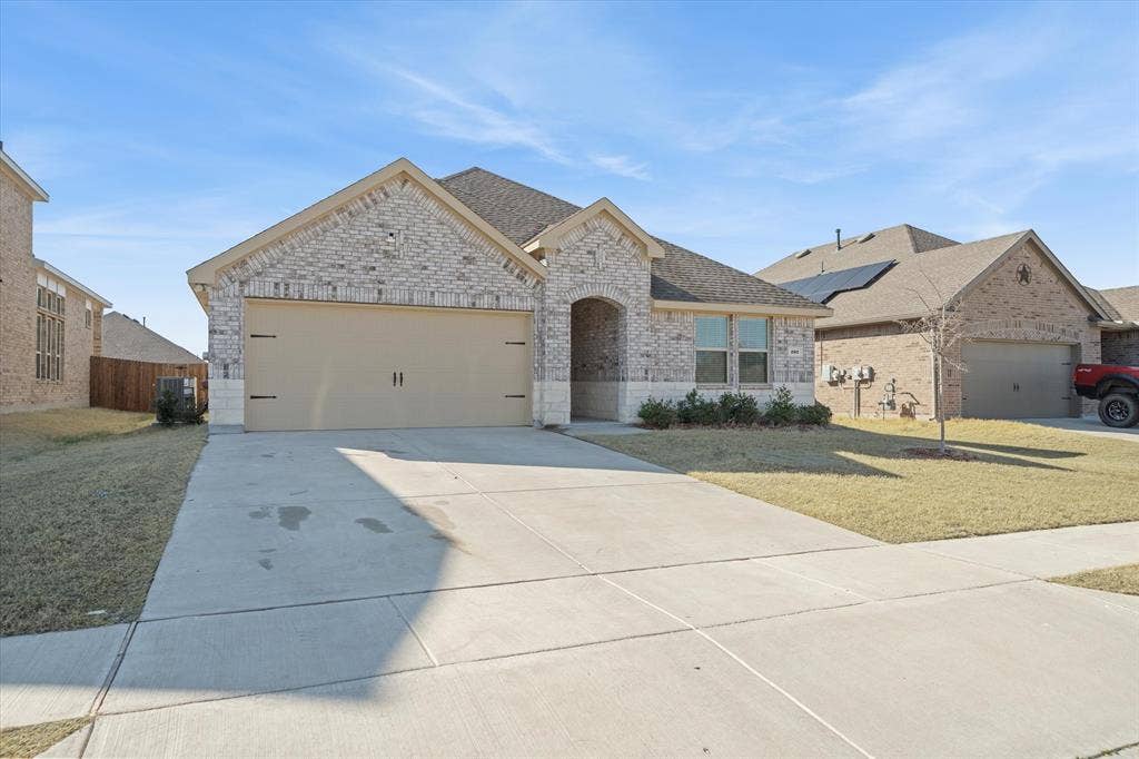 202 Buckboard Lane, Caddo Mills, TX 75135 Located at 202 Buckboard Lane in Caddo Mills, this single-story suburban residence presents strong curb appeal within a master-planned community. The front elevation showcases an attractive multi-tone brick and limestone facade beneath multi-gabled asphalt shingle roofing. A spacious double-car garage with carriage-style accents anchors the wide concrete driveway, bordered by a neat sidewalk and level lawn. An arched covered entryway welcomes visitors alongside large front windows, while privacy fencing stands between adjacent single-family properties beneath clear blue skies, offering a complete snapshot of modern neighborhood living.