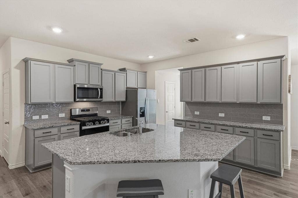 202 Buckboard Lane, Caddo Mills, TX 75135 Inside 202 Buckboard Ln, this open-concept kitchen features an expansive angled island fitted with speckled granite countertops, an undermount double-basin sink, and breakfast-bar seating. Sleek light-gray shaker cabinetry with dark hardware coordinates with a gray subway tile backsplash and durable light wood-look plank floors. Quality stainless-steel appliances include a gas range, mounted microwave, and French-door refrigerator, all illuminated by recessed LED ceiling lights. The thoughtful layout provides seamless flow to adjoining living areas, delivering an ideal culinary centerpiece for modern Texas living.
