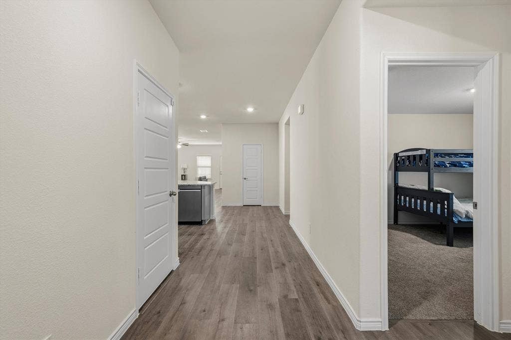 202 Buckboard Lane, Caddo Mills, TX 75135 Inside this home at 202 Buckboard Ln, a wide central hallway showcases durable, cool-toned wood-grain plank flooring paired with crisp white trim. Recessed LED lighting illuminates the path as it transitions seamlessly past a carpeted secondary bedroom on the right, which comfortably accommodates bunk bedding. Ahead, the corridor opens into an airy open-concept living space and kitchen centered around a stone-topped island with a stainless steel dishwasher, highlighting a thoughtfully designed single-story interior layout crafted for modern, functional everyday living.