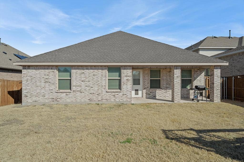 202 Buckboard Lane, Caddo Mills, TX 75135 The backyard of 202 Buckboard Ln features a private, level lawn fully enclosed by wooden privacy fencing. The single-story brick home showcases a hipped asphalt shingle roofline, multiple hung windows, and a pet-ready rear access door. An integrated covered patio, supported by matching brick pillars, provides a sheltered outdoor retreat complete with space for grilling and entertaining. Abundant natural sunlight bathes this quiet outdoor space, offering ideal privacy and open sky views within a welcoming Caddo Mills residential community.