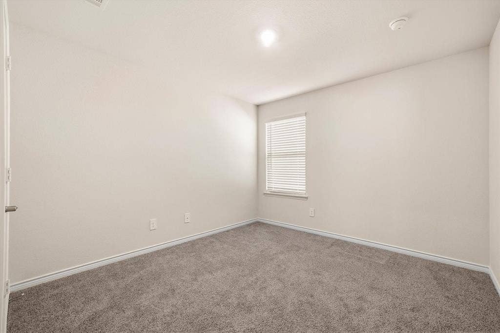 202 Buckboard Lane, Caddo Mills, TX 75135 Inside this move-in-ready residence at 202 Buckboard Ln in Caddo Mills, a secondary bedroom presents a versatile, well-lit retreat. The functional interior showcases soft, neutral-toned walls complemented by clean white baseboards and plush, heathered wall-to-wall carpeting underfoot. Natural daylight streams through a tall exterior window fitted with horizontal blinds, balanced by central overhead lighting and dedicated HVAC venting. Complete with modern electrical outlets and a standard entry door, this bright space easily adapts to a private guest suite, home office, or quiet personal haven.