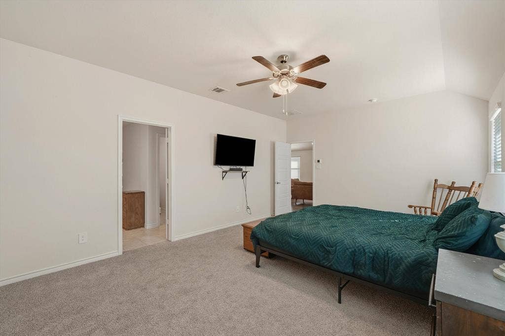 202 Buckboard Lane, Caddo Mills, TX 75135 A spacious primary bedroom suite at 202 Buckboard Ln offers a restful retreat featuring plush neutral carpeting, soft beige walls, and high ceilings with an integrated ceiling fan. Natural light pours in through side windows fitted with horizontal blinds, illuminating room for a king-size bed and sitting furniture. The functional layout provides immediate access to an attached en-suite bathroom with tiled floors as well as an open doorway leading back to the home's main living areas, delivering both everyday convenience and privacy.