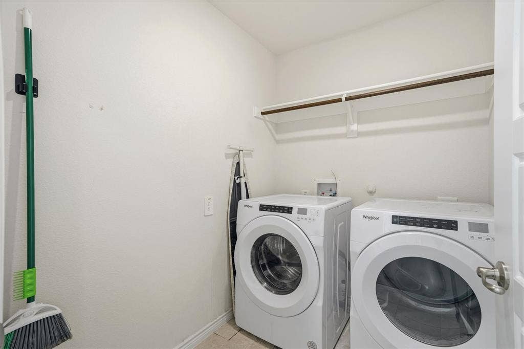 202 Buckboard Lane, Caddo Mills, TX 75135 A dedicated interior utility room at 202 Buckboard Ln provides efficient laundry functionality with neutral textured walls and durable ceramic tile flooring. The space is equipped with side-by-side white front-loading Whirlpool washer and dryer units beneath an overhead storage shelf with an integrated clothes hanging rod. Recessed plumbing connections, organized wall mounts for cleaning tools, and bright finishes make this practical laundry area a tidy, move-in ready convenience within the home.