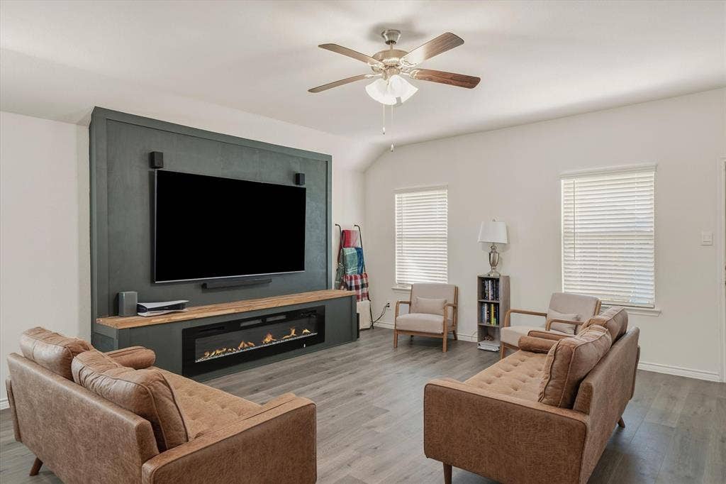202 Buckboard Lane, Caddo Mills, TX 75135 The living room at 202 Buckboard Ln presents a welcoming, open atmosphere defined by a dramatic vaulted ceiling and durable wide-plank, grey-toned wood-look flooring. A striking focal point anchors the space with a rich, dark matte green accent wall holding a mounted flat-screen television, natural wood mantle, and linear electric fireplace. Soft sunlight filters through two side windows dressed in horizontal blinds, illuminating the balanced seating arrangement and overhead brushed-nickel ceiling fan to create a stylish, functional everyday retreat in this Caddo Mills home.