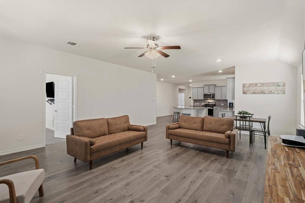 202 Buckboard Lane, Caddo Mills, TX 75135 This bright, open-concept interior at 202 Buckboard Ln showcases a seamlessly connected living, dining, and kitchen layout ideal for modern entertaining. Neutral walls complement continuous grey wood-grain luxury vinyl plank flooring, recessed lighting, and a brushed-nickel ceiling fan. The staged gathering room transitions effortlessly toward an intimate dining alcove and a well-appointed kitchen featuring grey shaker cabinetry, granite-look countertops, an eat-in peninsula, and stainless steel appliances. An adjacent doorway reveals a secondary carpeted living space, accentuating the flexible design of this inviting Caddo Mills home.