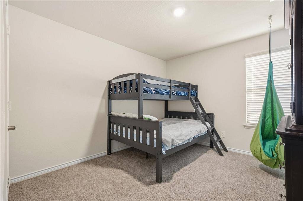 202 Buckboard Lane, Caddo Mills, TX 75135 Sunlight filters through a single-hung window dressed with horizontal blinds, illuminating a comfortable secondary bedroom at 202 Buckboard Ln. The functional layout features soft neutral walls, crisp white baseboards, and durable low-pile carpeting throughout. A dark gray twin-over-full bunk bed makes efficient use of the corner, while generous floor space accommodates a playful suspended pod swing and complementary bedroom furnishings. Positioned near a white interior panel door and bathed in flush-mount ceiling light, this adaptable bedroom offers practical comfort within this inviting Caddo Mills residence.