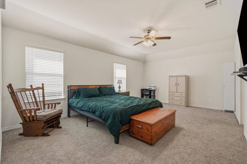 202 Buckboard Lane, Caddo Mills, TX 75135 A spacious bedroom interior at 202 Buckboard Ln showcases high tray ceilings, neutral off-white walls, and plush beige carpeting. Twin single-hung windows equipped with horizontal blinds provide ample natural lighting across the room, while a central five-blade wood ceiling fan circulates air efficiently. The generous floor plan accommodates a king-size bed frame, accent glider seating, a versatile work desk, and multiple freestanding armoires, delivering a restful and highly functional living retreat inside this comfortable Caddo Mills home.