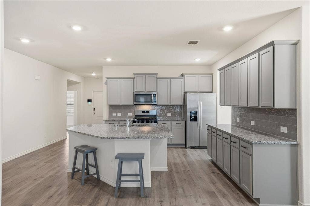 202 Buckboard Lane, Caddo Mills, TX 75135 Inside 202 Buckboard Ln, this contemporary kitchen features a seamless open-concept design complete with durable grey wood-look plank flooring. The space is anchored by an angled prep island with breakfast bar seating and an integrated sink, highlighted by speckled granite countertops and glossy subway tile backsplashes. Stainless steel appliances, including a gas range and side-by-side refrigerator, complement the light grey Shaker cabinetry with modern black hardware. Recessed ceiling lights illuminate the inviting layout, providing smooth sightlines into the adjoining living spaces of this Caddo Mills residence.
