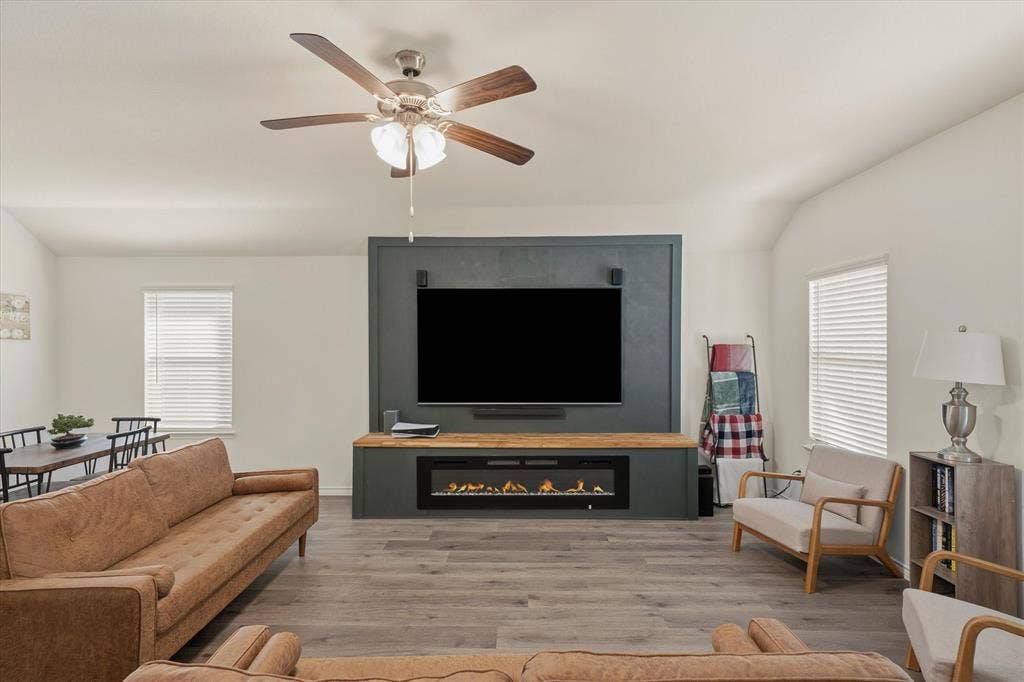 202 Buckboard Lane, Caddo Mills, TX 75135 The spacious living room at 202 Buckboard Ln showcases an inviting open-concept layout centered around a custom dark-teal media accent wall. A linear electric fireplace set beneath a natural wood mantel serves as a cozy focal point below the wall-mounted television. Natural light filters through dual windows flank the fireplace, illuminating wide-plank wood-look flooring and soaring vaulted ceilings equipped with a modern ceiling fan. This comfortable interior effortlessly connects living and dining spaces, offering a warm and stylish everyday gathering environment.