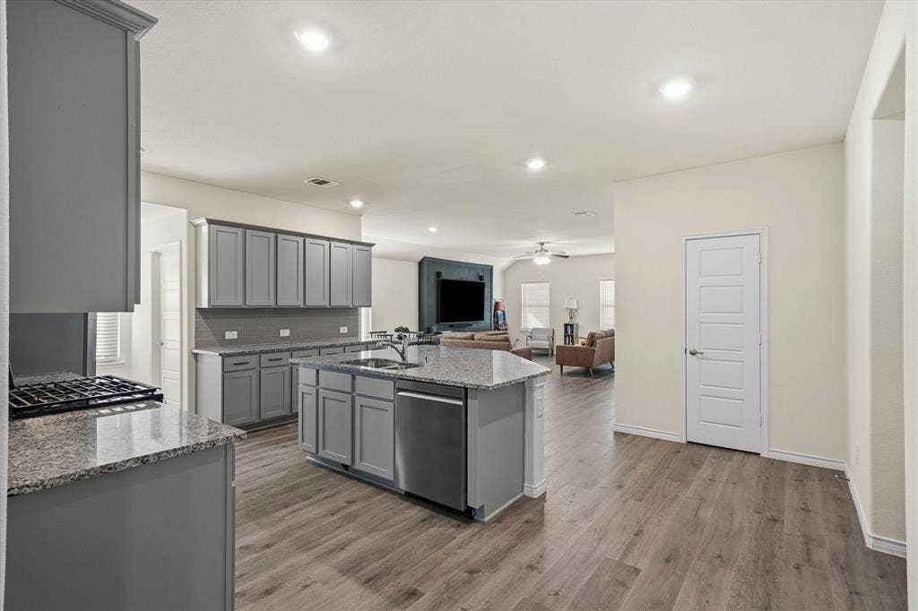 202 Buckboard Lane, Caddo Mills, TX 75135 This open-concept interior at 202 Buckboard Ln showcases seamless suburban living, connecting a modern kitchen to the primary living space. Light grey shaker cabinetry, granite countertops, and a subway-tile backsplash frame the culinary area, centered by an expansive prep island with an undermount double sink and breakfast bar seating. Continuous wood-look luxury vinyl plank flooring unites the entire main level beneath recessed lighting, leading directly to a welcoming living room complete with built-in cabinetry, an entertainment wall, and bright windows fitted with horizontal blinds.