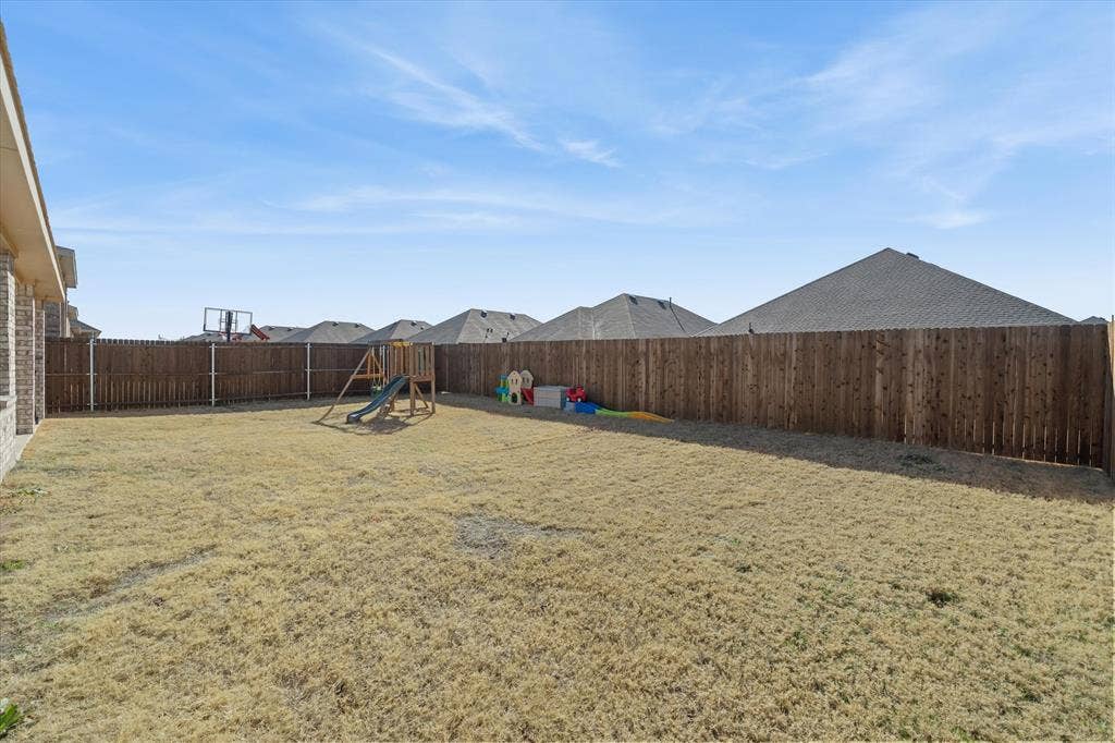 202 Buckboard Lane, Caddo Mills, TX 75135 Expansive fully fenced backyard at 202 Buckboard Ln in Caddo Mills, offering a private outdoor retreat within a welcoming residential subdivision. The level yard is completely secured by a tall wood privacy fence with metal reinforcement posts, bordering neighbor single-family homes beyond. Ideal for play and recreation, the open lawn comfortably accommodates outdoor playsets and toys alongside the brick exterior of the primary home. Bright daytime sunlight highlights the generous outdoor footprint, presenting boundless potential for custom landscaping, pets, entertaining, and outdoor living.