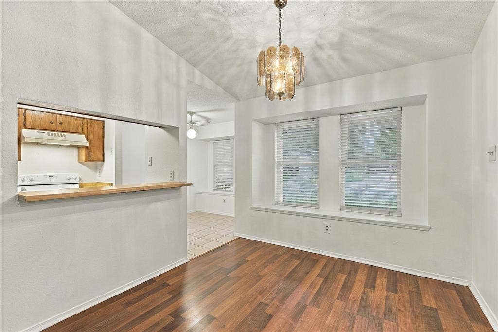 844 Woodrock Drive, Dallas, TX 75217 Vaulted ceiling with an ornate chandelier, wood-finish flooring, two large windows with blinds, and a pass-through opening to the kitchen area with wood cabinetry and a white range