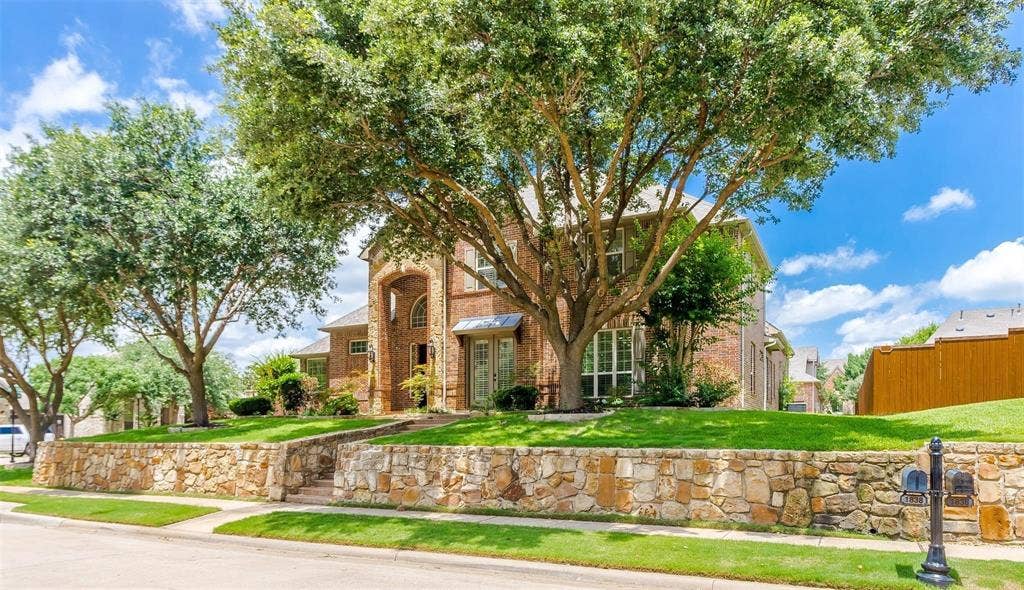 1838 Olney Drive, Allen, TX 75013 Traditional brick residence featuring an arched portico entry, mature trees, manicured lawn, and a stone retaining wall with integrated steps
