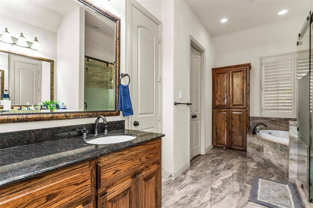 1838 Olney Drive, Allen, TX 75013 Vanity with a dark countertop and undermount sink, framed mirror, built-in wooden cabinet, soaking tub with tile surround, and plantation shutters