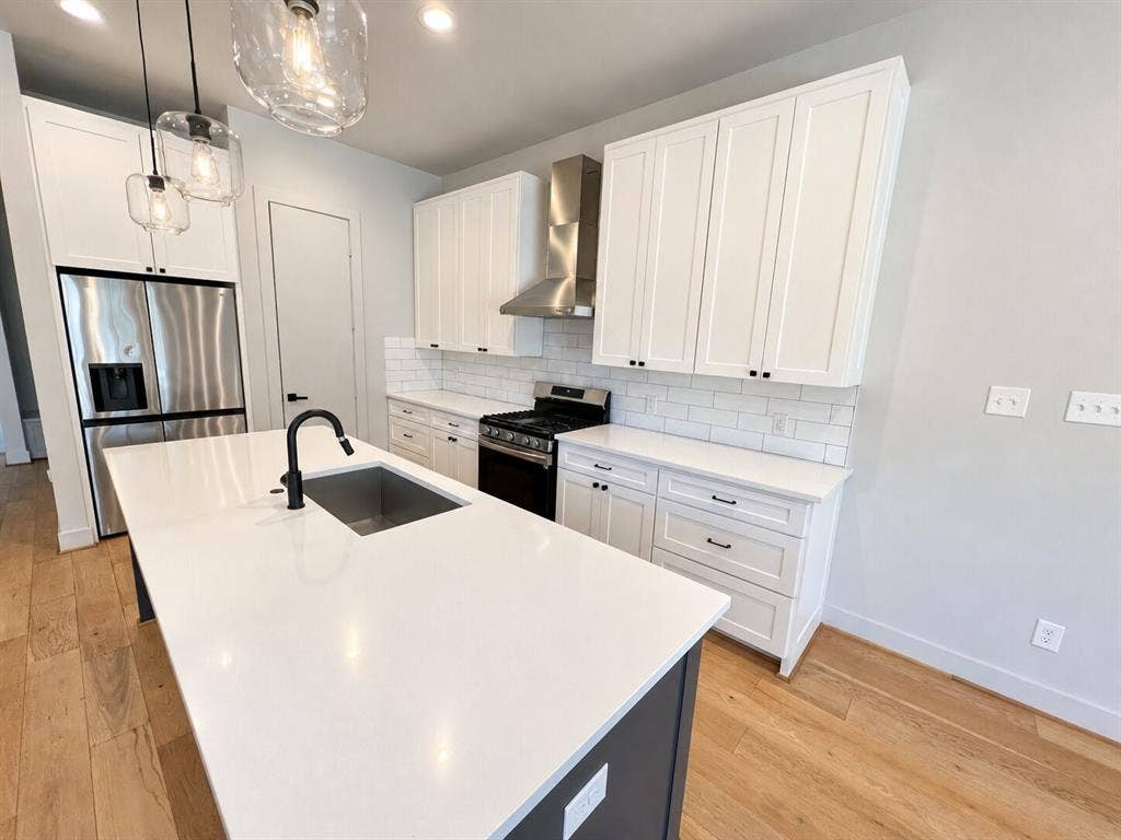 Contemporary kitchen featuring a large center island with an undermount sink, stainless steel appliances, white shaker cabinetry, white subway tile backsplash, and light wood-finish flooring