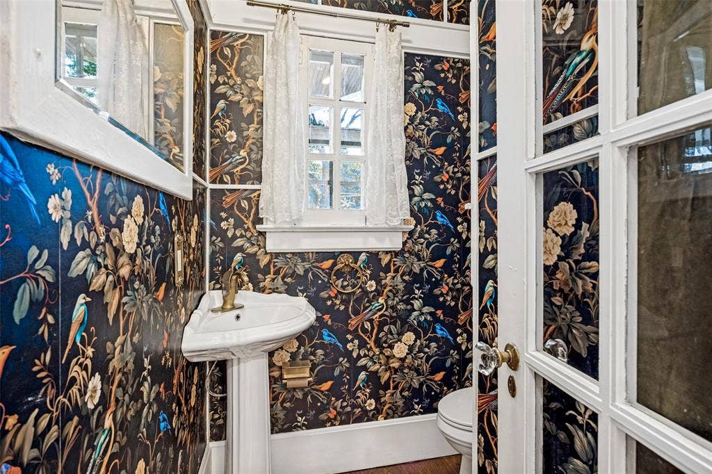 320 E 5th Street, Dallas, TX 75203 Powder room featuring bold botanical wallcovering in navy, gold, and blue tones, pedestal sink with chrome hardware, white painted window casement with multiple panes, and wood-finish flooring